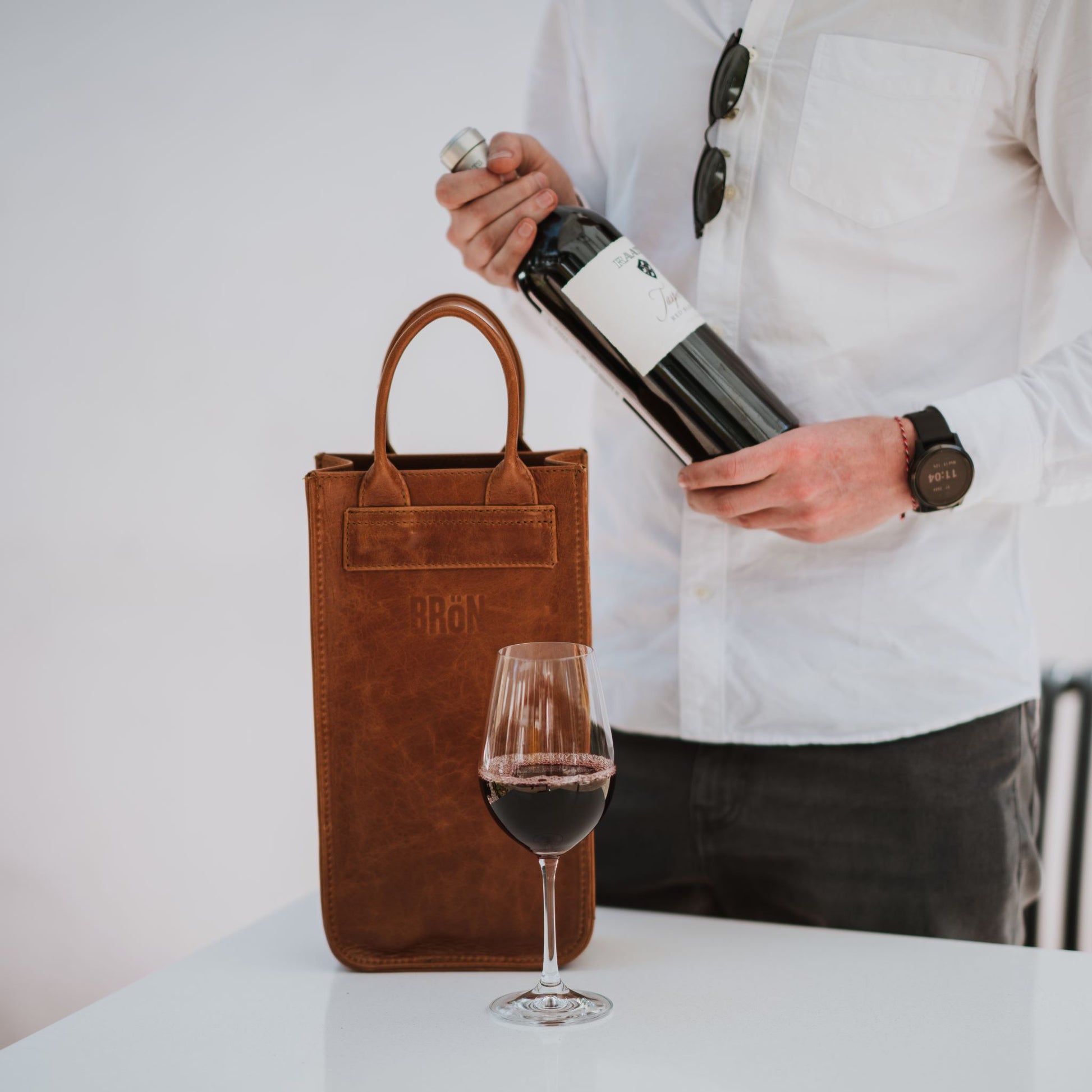 Front view of the leather wine carrier, showcasing the handles, a glass of wine and a bottle of wine.