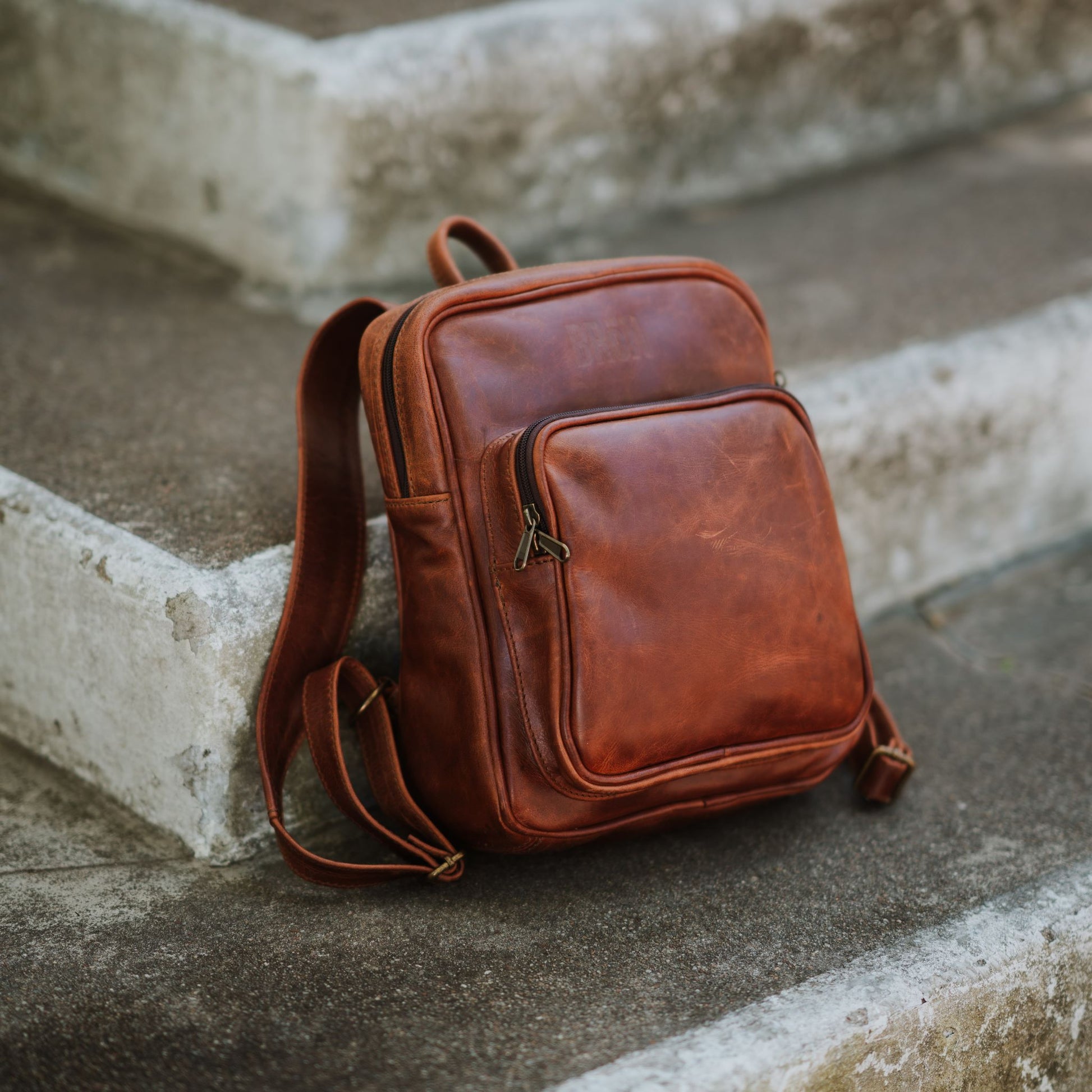 Front and side view of the small leather backpack, showcasing the front zip pocket, shoulder straps and handle.